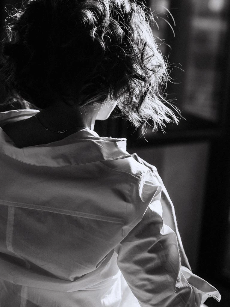 A reader in a sunlit Brooklyn apartment, dressed in cashmere, beside a stack of books.